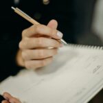 A close-up of a hand holding a pen and writing in a spiral notebook.